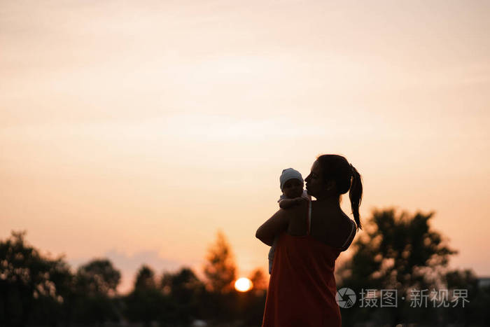 日落剪影年轻的母亲抱着她的男婴在城市公园站在夕阳和鲜艳的橙色天空