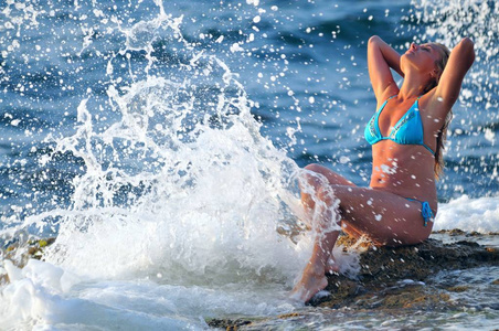 身穿蓝色比基尼的金发女子坐在岩石上享受着海浪的滴滴