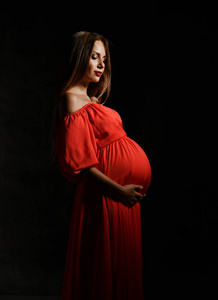 年轻漂亮的孕妇穿着红色的衣服站着，抱着肚子微笑着