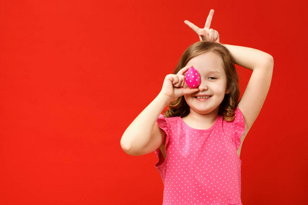 快乐的手指孩子做兔子耳朵，拿着一个粉红色的复活节彩蛋。红底小女孩的画像