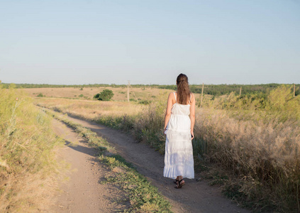 沿着草地走一个年轻的女孩,穿着白衣服,独自在田野里,沿着小路走着.