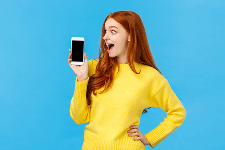 哇看看这个应用程序。又好笑又好奇，兴奋的红发女孩看着智能手机显示屏，显示手机屏幕购物网站或应用程序，站在蓝色背景下