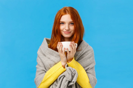 可爱浪漫帅气的红发女，戴着灰色围巾，暖和起来，喝着热茶，端着杯咖啡，微笑着的相机高兴而放松，站在蓝色的背景下很高兴