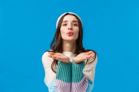 性感的深色女孩在冬季毛衣,帽子,拉近相机折叠嘴唇和举起手臂,吹气吻