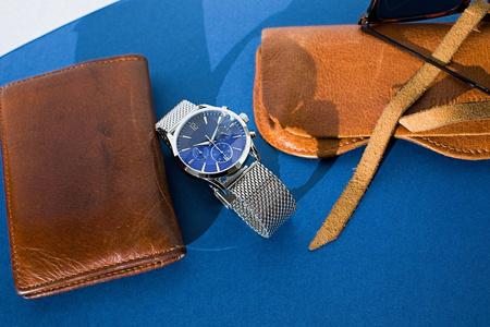皮包，带金属手镯的手表，墨镜和蓝色背景的笔记本电脑。男士配件。俯视图