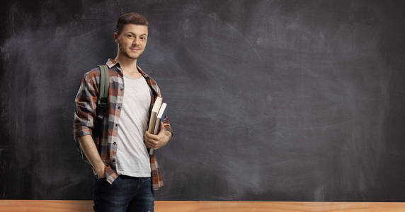 Male student in front of a blackboard holding books 