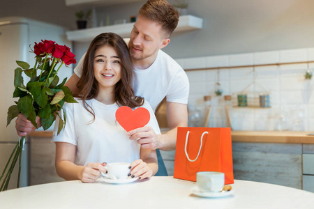 年轻帅气的男人在情人节早上在厨房喝咖啡，送上玫瑰和礼物给心爱的妻子一个惊喜，浪漫纪念日快乐
