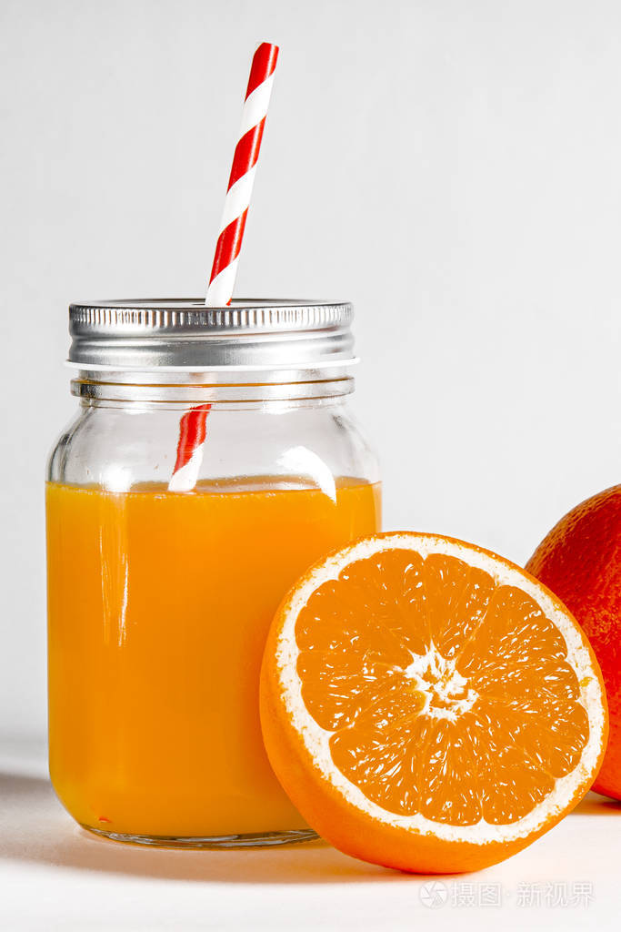 一个装着鲜榨橙汁的玻璃罐立在白色的背景上旁边是新鲜的橘子