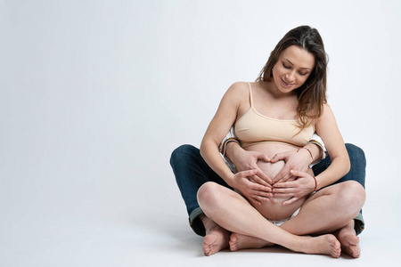 年轻人抱着怀孕妻子的肚子做心脏。孕妇和慈爱的丈夫在家抱着肚子。手心由可爱的夫妇在怀肚子上。复制空间，隔离