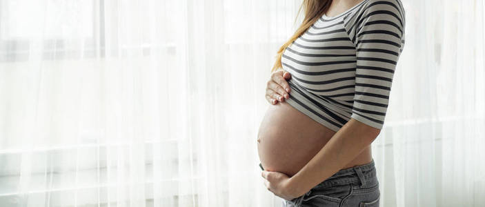幸福的大肚子孕妇在窗边。怀孕和家庭的概念