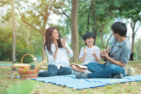 快乐的亚洲家庭在公共场合有闲暇帕克，神父弹着吉他，母子俩一起拍手，享受着幸福的脸庞。