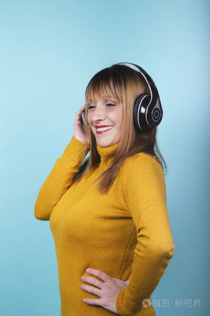 身穿黄色套头衫的女士戴着耳机微笑着听音乐，在蓝色背景下微笑。孤立的图像。