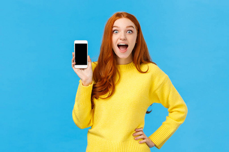 惊讶和感动，兴奋的红发女黄毛衣介绍新的应用程序，显示智能手机显示屏，微笑迷住张嘴逗乐，看相机，蓝色背景