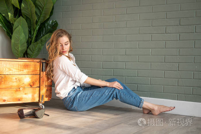 年轻漂亮的女人光着脚坐在木箱和绿色植物旁边的地板上