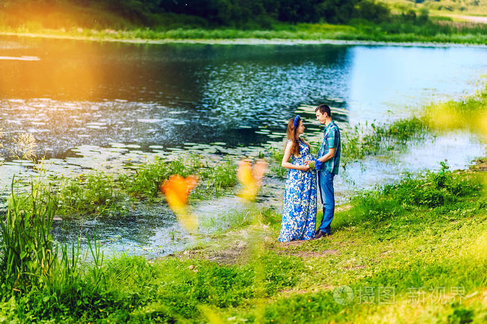 年轻幸福的怀孕夫妇在日落时在河边互相凝视夏天新家庭在河边风景优美
