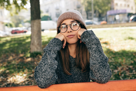 悲伤的年轻女子看着镜头，戴着时髦的透明眼镜，毛衣和帽子。美丽的女性坐在公园的长椅上休息。