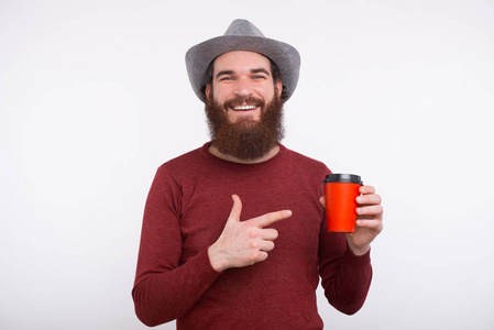 在白墙附近，一个留着胡子的男人指着一个盛着热饮戴着帽子微笑的杯子。