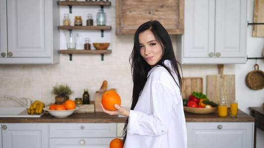 年轻的黑发女人拿着橘子在厨房微笑着。