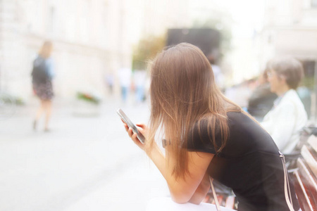 一个年轻漂亮的女孩正在看一部智能手机。一个女人坐在一座古城中心的长椅上，一片模糊。现代青年的生活方式与消遣