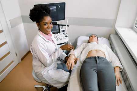 微笑的年轻非洲女医生用超声波探头在躺在沙发上的年轻白人女性患者的手腕上