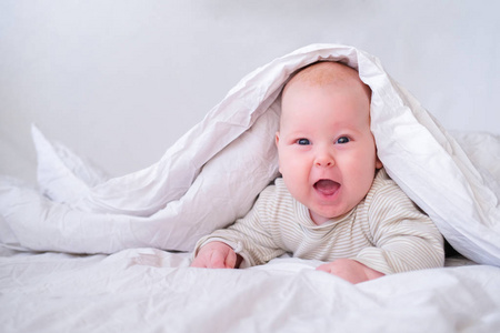 卧室里可爱的微笑着的小男孩躺在白色毯子下。新生儿在床上休息。家人早上在家。新生婴儿