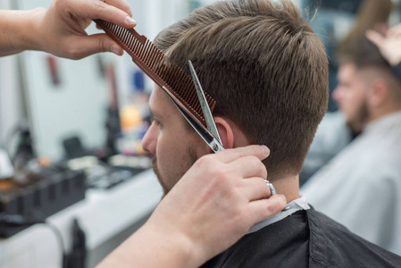 理发师师傅给一个留胡子的年轻人理发。理发店的专业理发师用剪刀的手的特写。