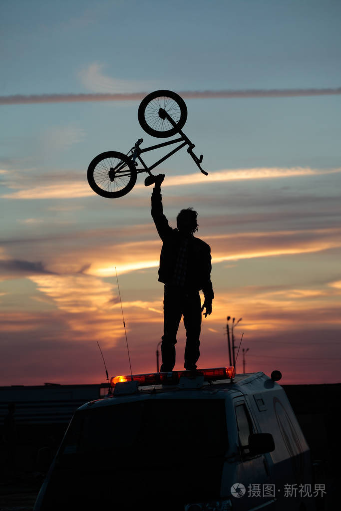 一个骑自行车的人在日落的背景下举着自行车站在一辆汽车旁边