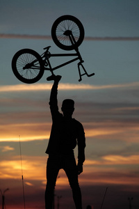 一个骑自行车的人在日落的背景下举着自行车，站在一辆汽车旁边