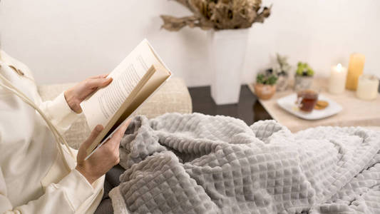 放松概念女人坐在一个现代室内的沙发上，腿上放着毯子，手里拿着一本纸质书