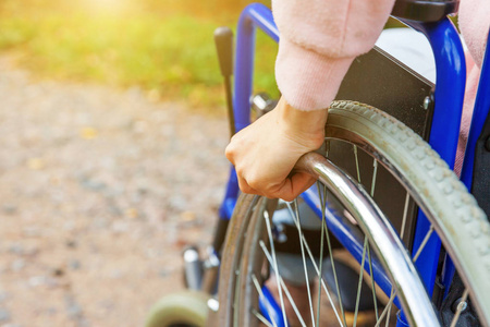 手残疾妇女坐在轮椅轮上，在医院公园的路上等待病人服务。无法辨认的瘫痪女孩坐在残疾人专用椅上。康复概念。