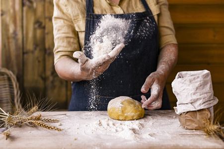 女人的手，面粉和面团。悬浮在一个面团和面粉的框架里。一个穿着围裙的女人正在准备面团，准备在家里烤。乡村风格照片。木桌，麦穗和面粉