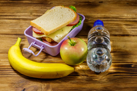 午餐盒，有三明治一瓶水香蕉和苹果放在一张木桌上