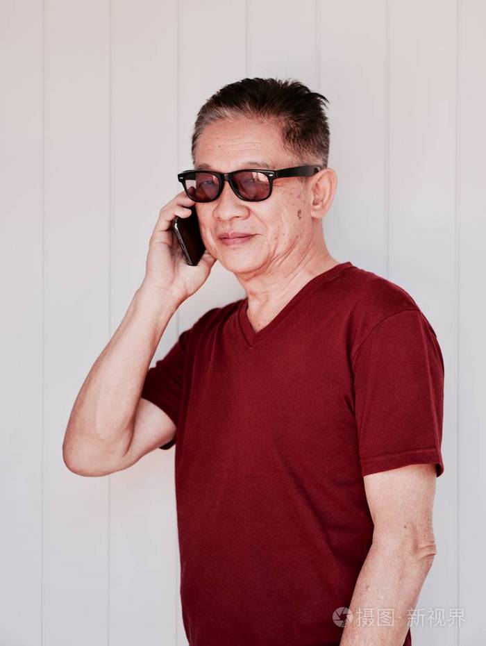 一位老人穿着红色t恤戴着墨镜用智能手机站在白墙前