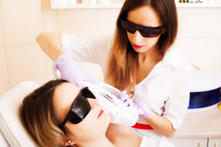 美容师在美容院为年轻女性面部进行激光脱毛治疗。