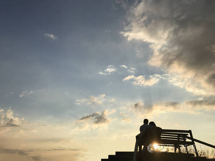 一男一女坐在长椅上对着夕阳两个恋人拥抱凝视远方一对情侣的剪影