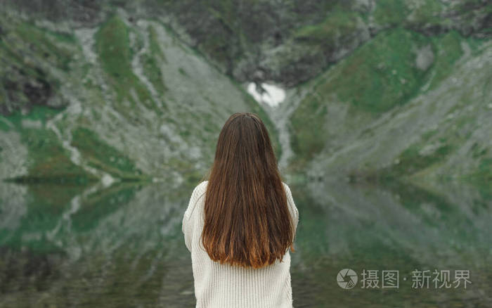 一个穿着毛衣的黑发女孩站在一个湖水的山上从后面