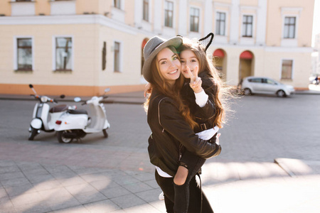 一个戴着灰色帽子的迷人女人背着黑发俏皮女孩在街上摆姿势.照片