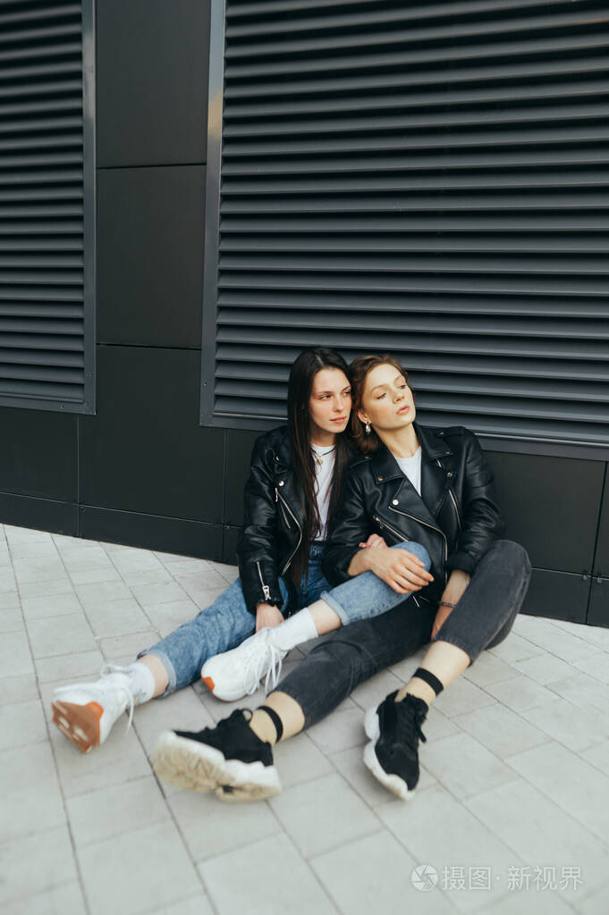 两个时尚女孩穿着休闲服穿着皮夹克坐在地上靠着一堵深色的墙看着离开