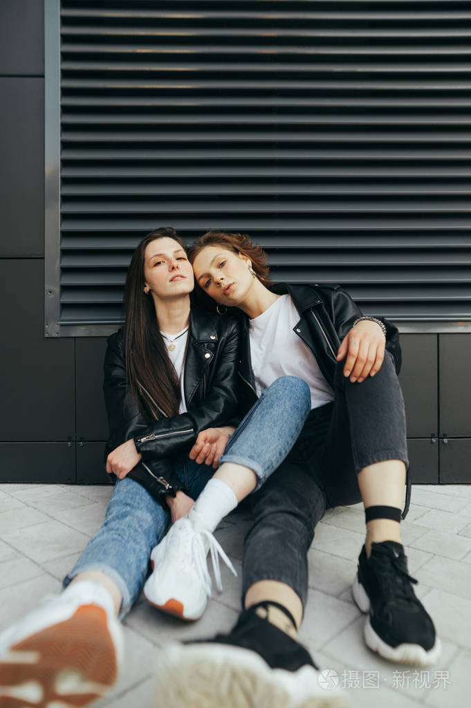 两个时髦女孩坐在深色背景上穿着深色衣服拥抱并摆姿势相机肖像两个穿