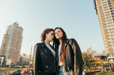 两个穿着街头服装的女孩站在城市景观背景上的草地上，对着镜头摆姿势。街上穿着皮夹克的迷人模特的画像。
