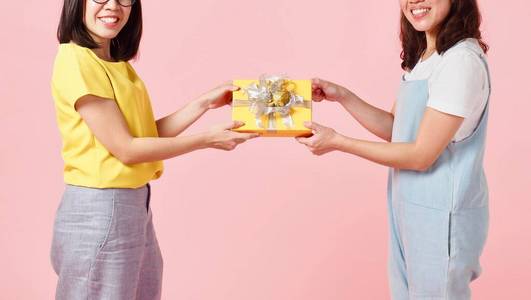 一位戴着眼镜的亚洲年轻女子把一个黄色的礼物盒送给一位面带微笑穿着蓝色牛仔裤粉色底色的女士