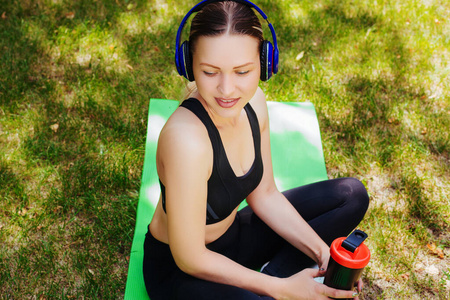 穿着运动服的漂亮女孩坐在公园里的绿色垫子上，手里拿着一杯水。戴着耳机做瑜伽。运动时听音乐