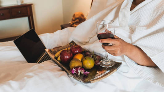 图为游客端着酒,用过的笔记本电脑,在床上吃水果,健康食品概念照片