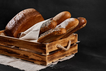 各种各样的新鲜出炉的面包放在一个纸袋里，木箱放在一个麻袋上，背景是黑色的，有复印空间。特写