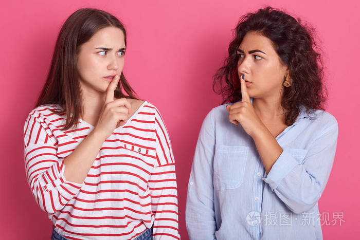 两个美丽的黑发女人表情严肃穿着休闲衬衫手指放在嘴唇上有着密谋的