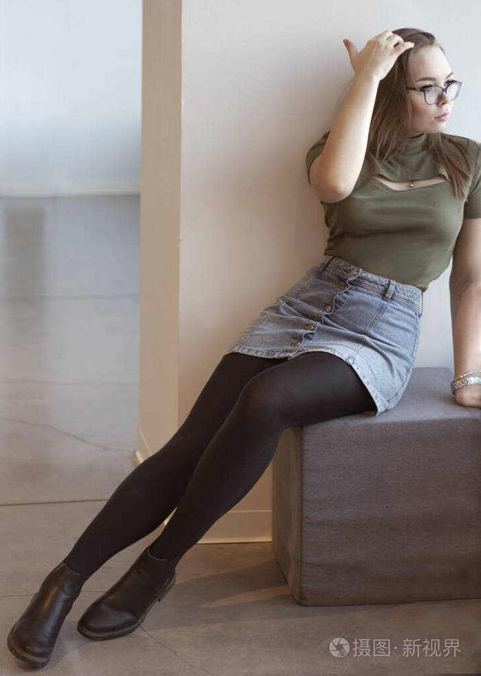 一个白人女孩坐在一张脚凳上两腿伸开向窗外望去