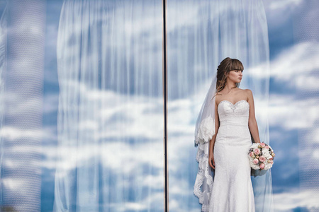 华丽奢华时尚的新娘身着白色礼服，手拿花束，在云天背景下的全景窗旁。