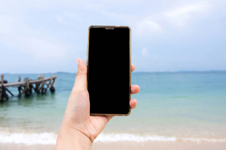 女性手持智能手机与海滩背景。剪辑路径显示屏幕包括在此图像中。