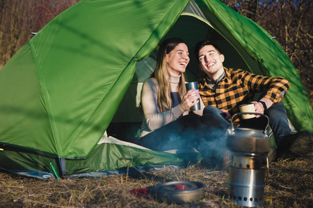 一对幸福的小情侣，微笑着，在篝火旁的帐篷里喝茶喝咖啡。青少年家庭户外活动