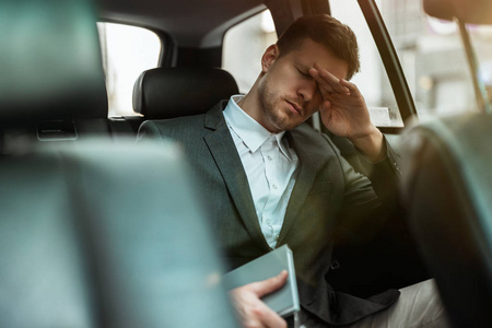 一个成功的商人手里拿着计划者，坐在汽车后座上与合作伙伴见面时，看上去筋疲力尽，一心多用的概念